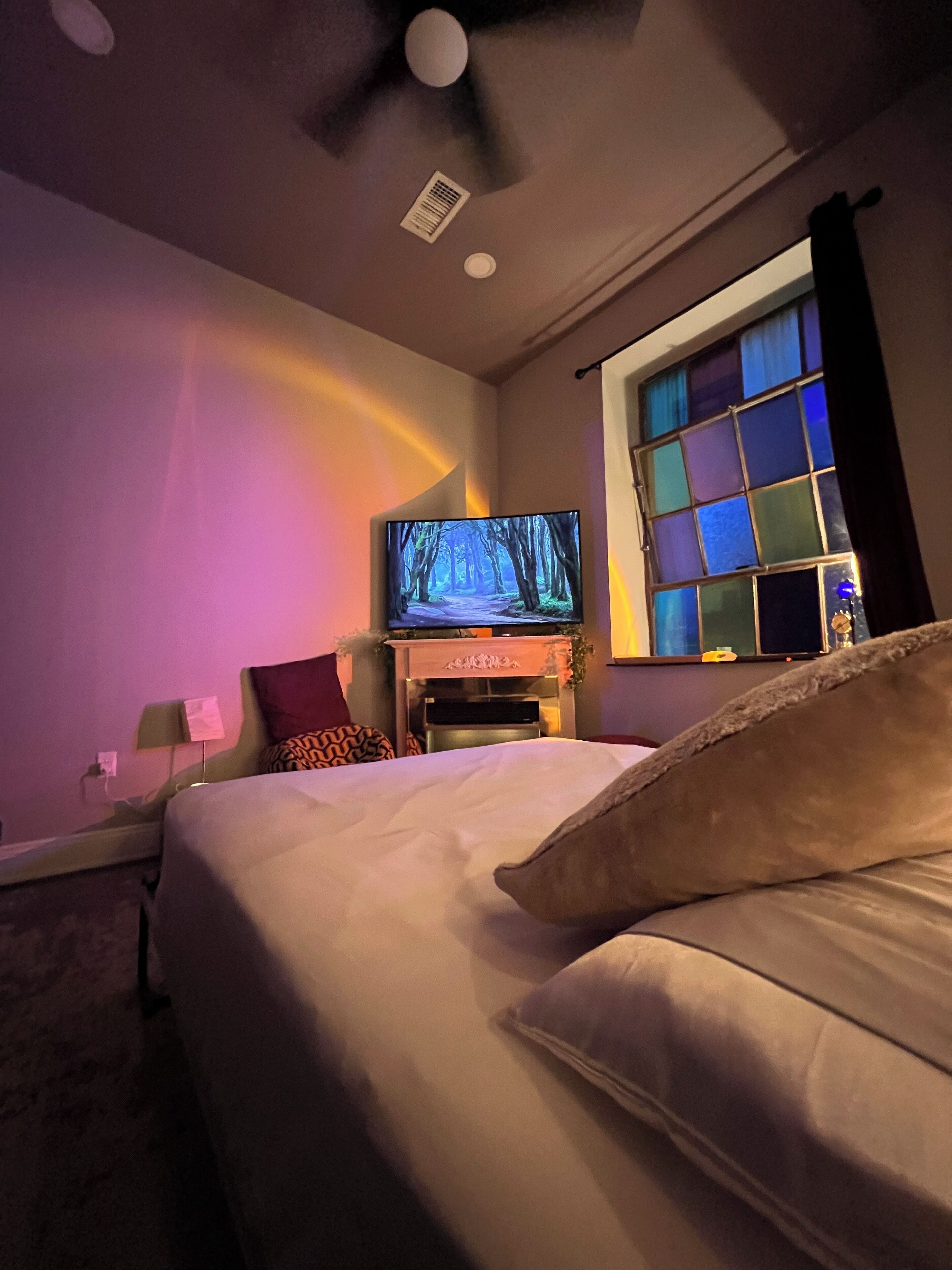A hotel room with a bed, a television, a window with a view of the night sky, and a ceiling fan.