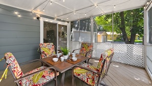 A covered patio with a table set for four, surrounded by colorful chairs, string lights, and a view of the outdoors. - Urban Nest | Garage, Sunroom & Fenced Yard (Fort Collins)