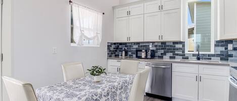 Cocina moderna con armarios blancos, salpicadero de azulejos negros y grises, y un comedor con mantel y sillas estampadas.