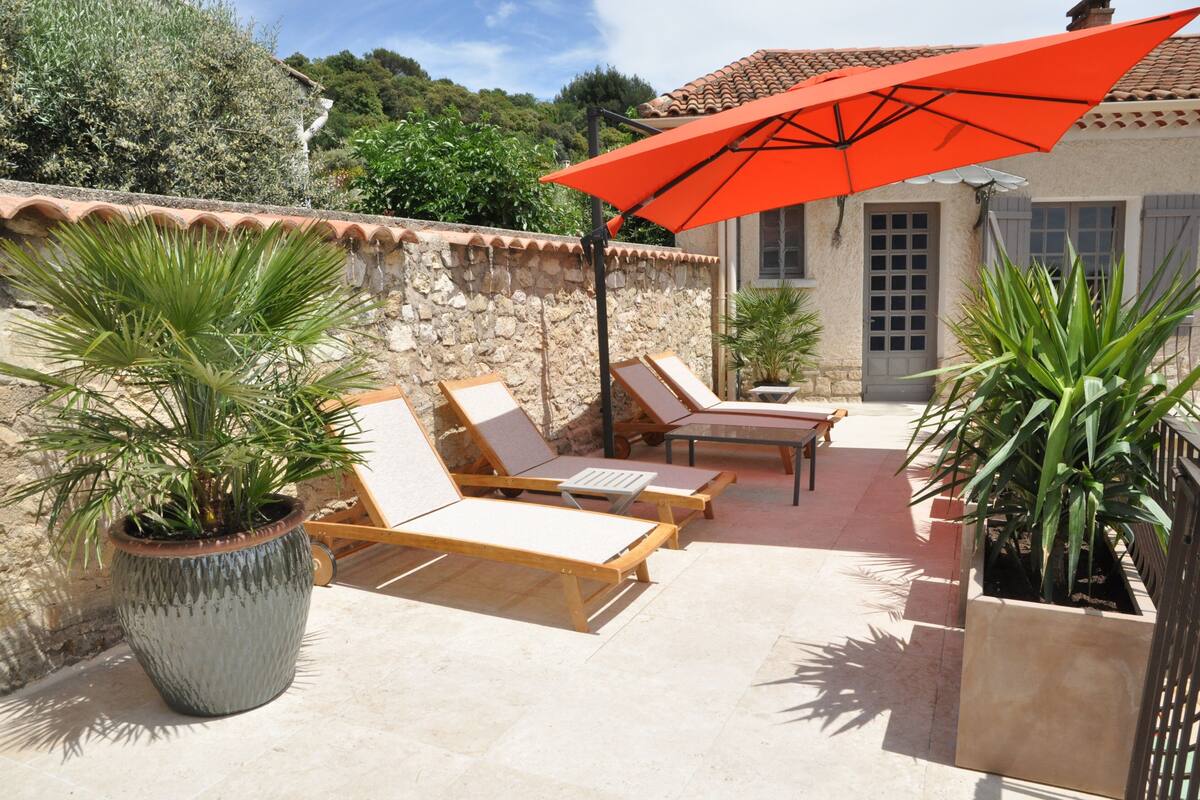 A sunlit patio with lounge chairs and a red umbrella, surrounded by stone walls and potted plants.