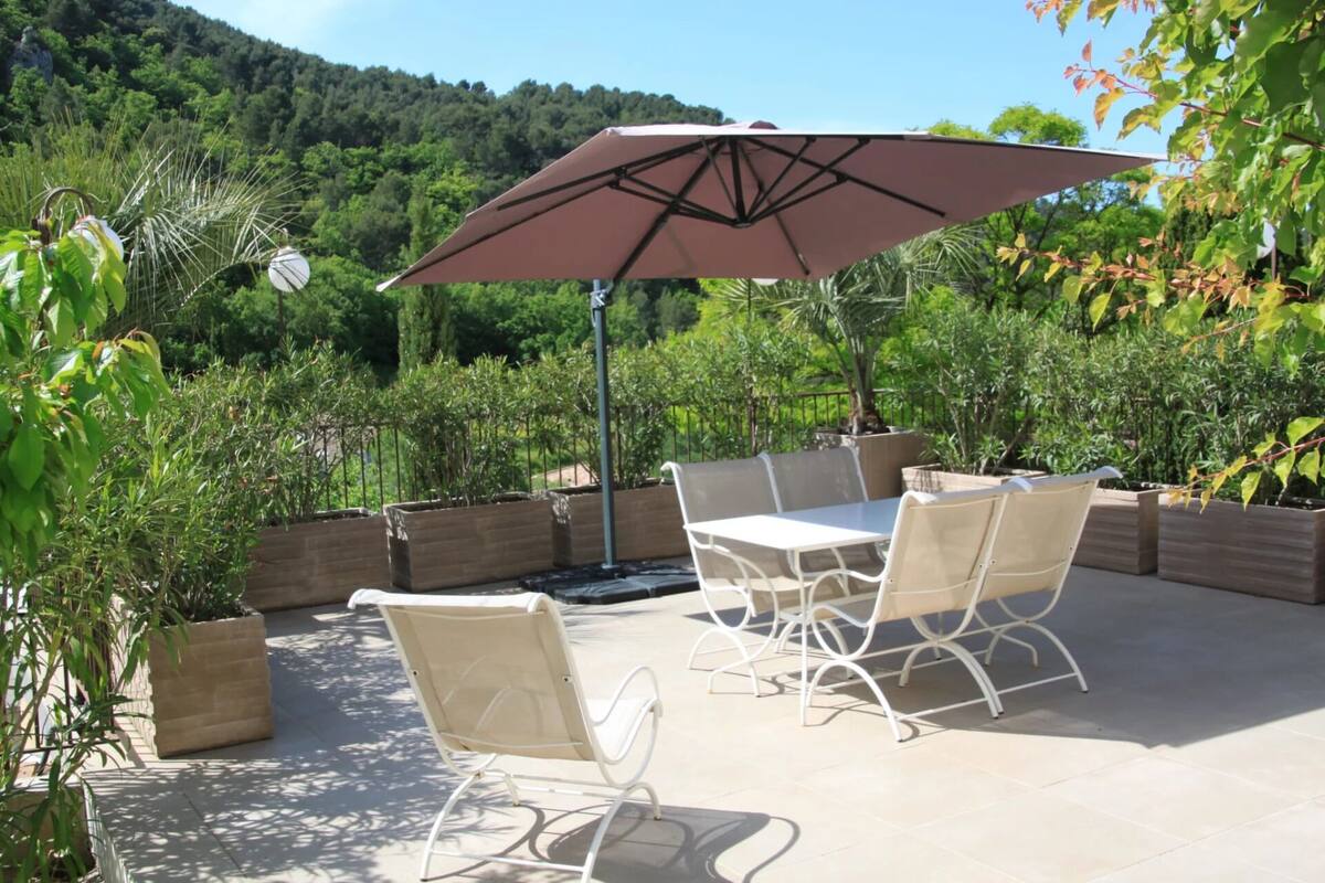 An outdoor patio with a table and chairs, surrounded by planters and a large umbrella.