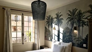 A bedroom with a large bed, a window with curtains, a wall mural of palm trees, and a hanging light fixture. - Les Terrasses de la Roque Alric (La Roque-Alric)