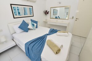 Ein ordentlich bezogenes Bett mit blauem Handtuch und zusammengerolltem Handtuch, ein weißer Nachttisch mit Lampe, ein gerahmtes abstraktes Gemälde, ein Spiegel und eine weiße Tür.