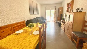 Un comedor con mantel a cuadros amarillo, sillas de madera y un banco. Hay un ventilador de techo, un televisor y un armario con cajones.