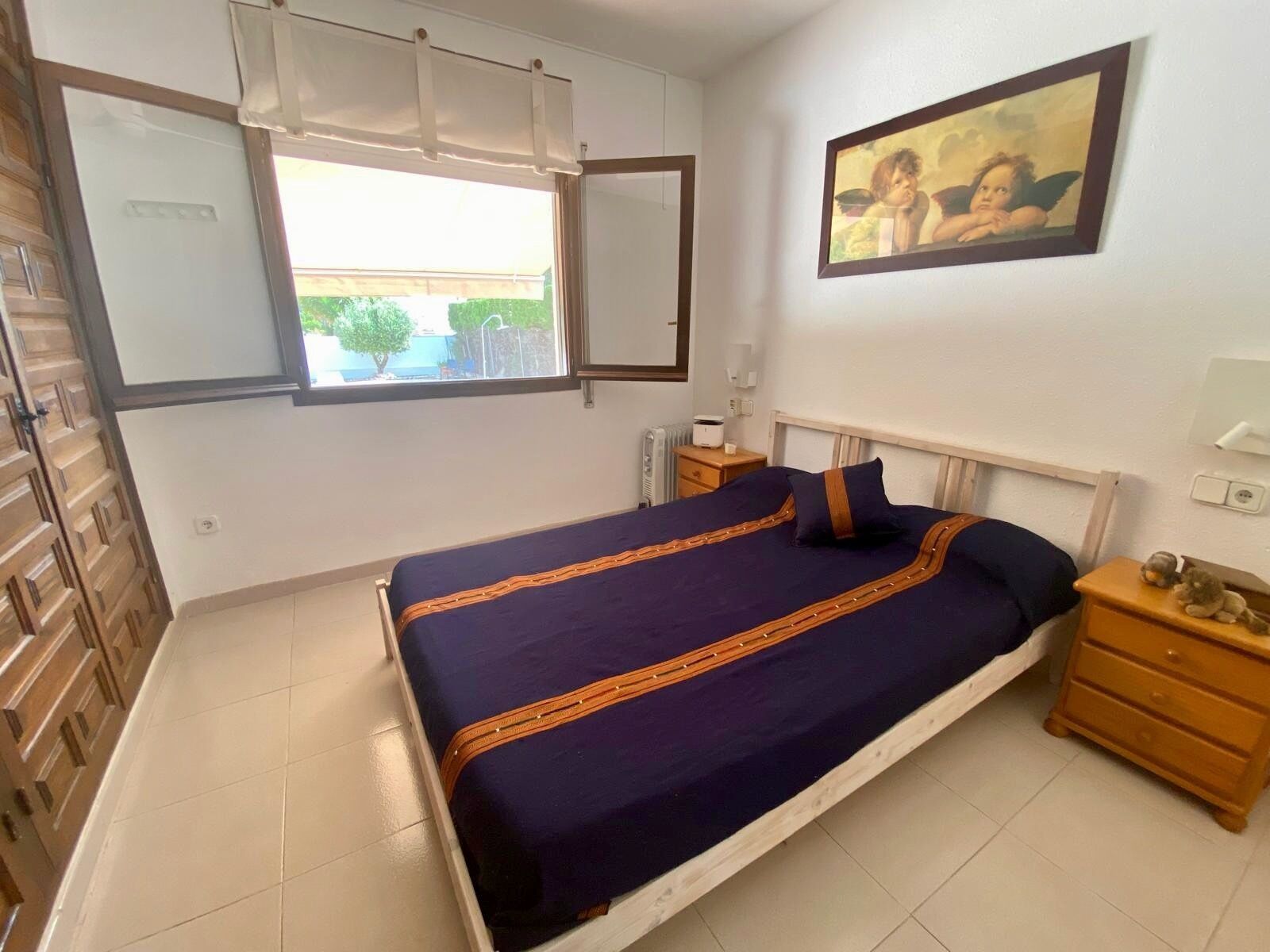 A bedroom with a bed, wooden nightstands, a window with blinds, a radiator, and a painting on the wall.