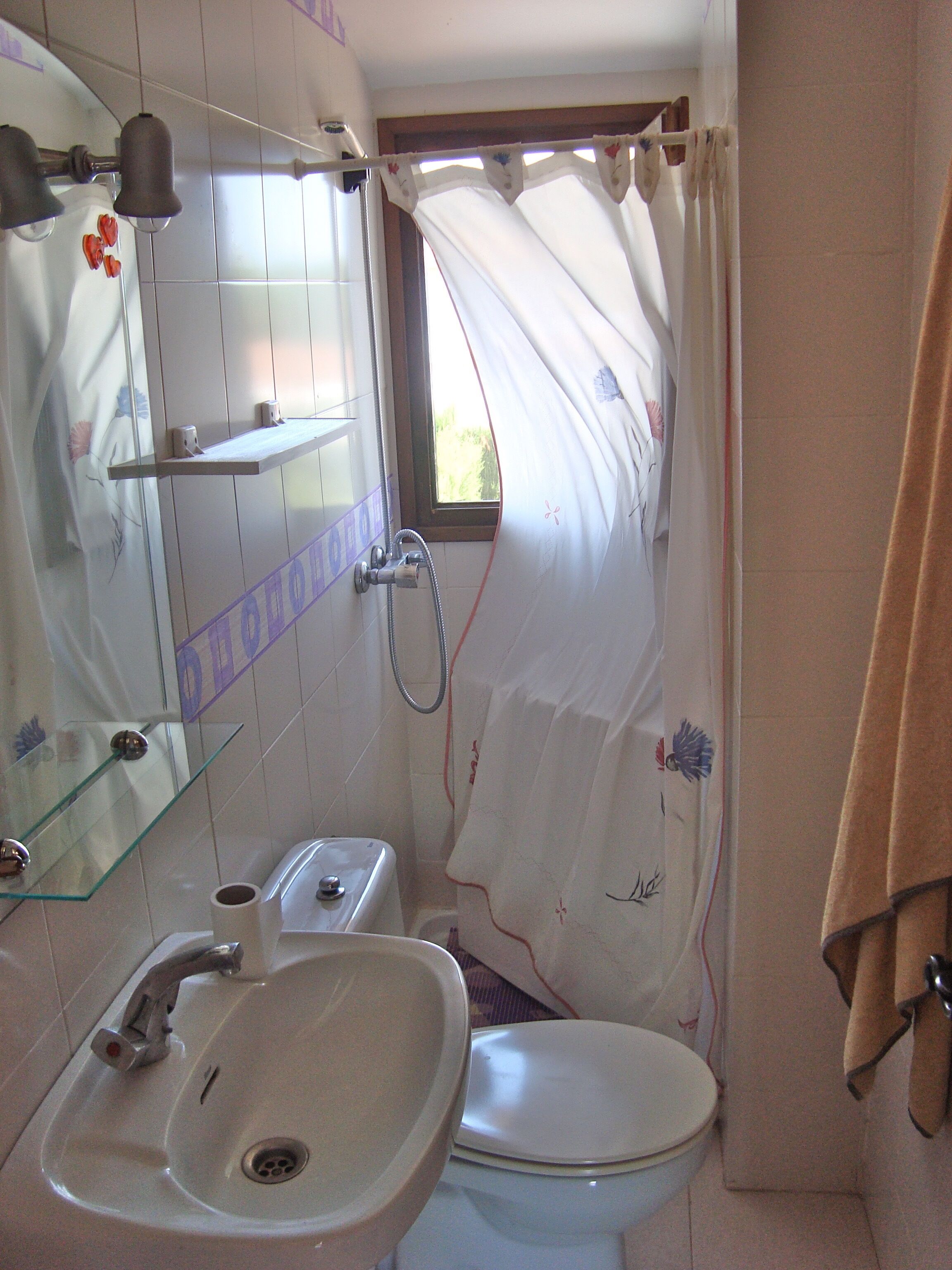Une salle de bain avec un lavabo, des toilettes et une douche. Il y a une fenêtre avec un rideau, et une serviette est accrochée au mur.