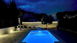 Una piscina exterior con borde iluminado en azul, rodeada de una zona pavimentada con sillas y una mesita.