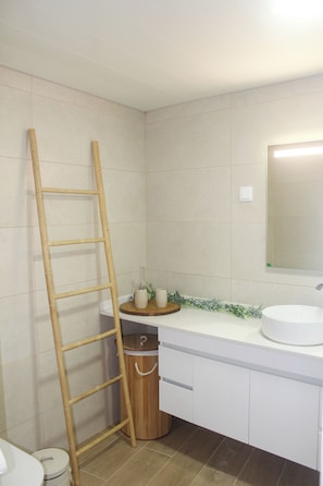 Een badkamer met een houten ladder tegen de muur, een toilet, een wastafel met spiegel en een kleine prullenbak.