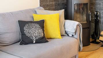 Un canapé gris avec trois coussins, un coussin noir orné d’un motif d’arbre et un coussin jaune. Un plaid gris est posé sur le canapé. On aperçoit un poêle à bois noir à l’arrière-plan.
