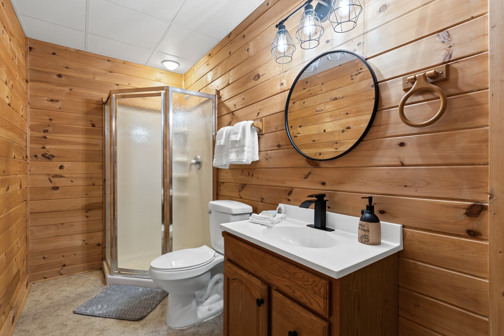 A wooden-paneled bathroom with a glass shower enclosure, a toilet, a sink with a soap dispenser, and a mirror.