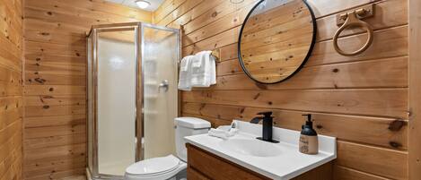 A wooden-paneled bathroom with a glass shower enclosure, a toilet, a sink with a soap dispenser, and a mirror.