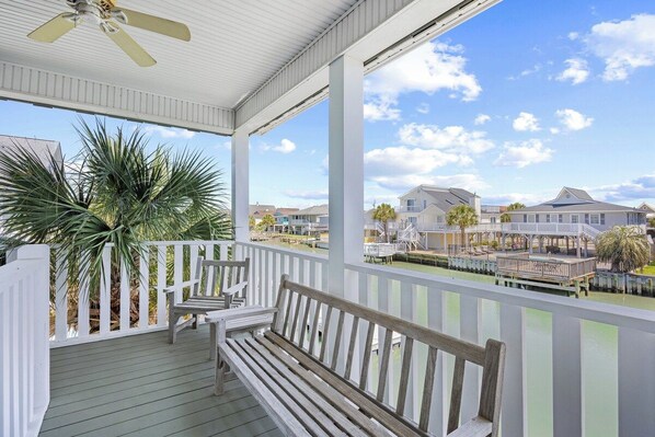 A covered porch with a ceiling fan, wooden benches, and a view of a residential area with houses and a dock.