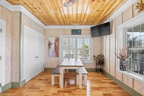 A room with a wooden ceiling, a large window, a table with benches, and a TV mounted on the wall.