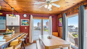 A kitchen with a wooden ceiling, a dining area with a table and chairs, and a view of buildings through large windows.