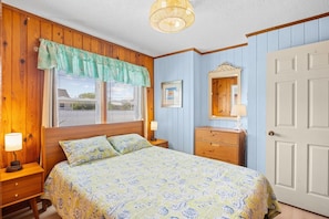 Een slaapkamer met een bed, nachtkastje, dressoir, raam met zonwering en een deur.
