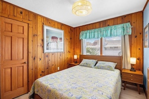 Een slaapkamer met een houten lambrisering, een bed met een geborduurde spreideken, een nachtkastje met een lamp en een raam met jaloezieën.