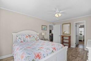A bedroom with a bed, a ceiling fan, a bedside table with a lamp, and a door leading to another room.