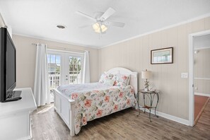 A bedroom with a bed, a ceiling fan, a TV, a window with curtains, and a small table with a lamp.