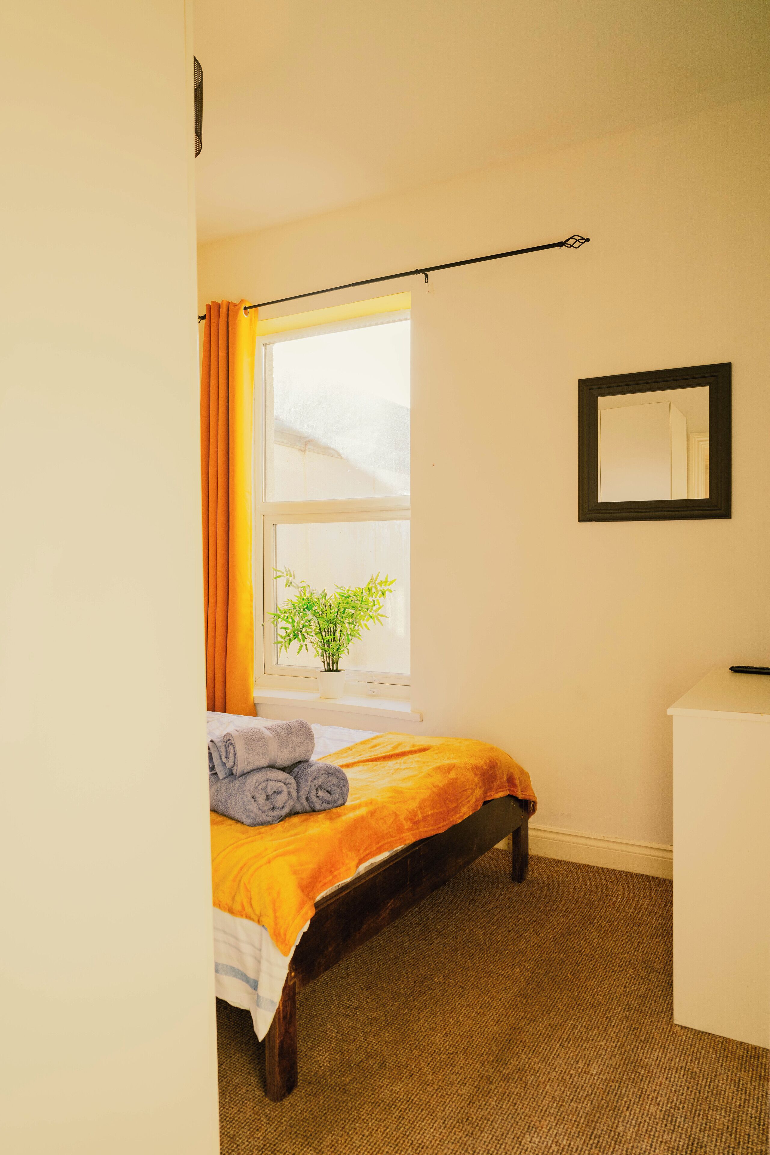 A bedroom with a bed, a window with curtains, a bedside plant, and a mirror on the wall.