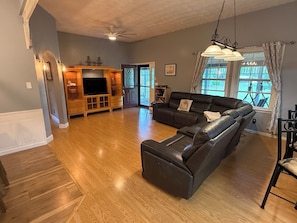 Un salon spacieux avec un grand canapé d’angle, un téléviseur à écran plat, un meuble de rangement en bois et une grande fenêtre avec des rideaux.