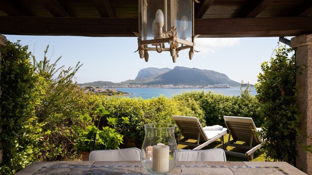 A table with a glass candle holder and a chair, overlooking a scenic view of a coastal area with mountains and a body of water. - Casa Acqua e Pietra by Wonderful Italy (Golfo Aranci)