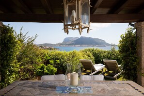 A table with a glass candle holder and a chair, overlooking a scenic view of a coastal area with mountains and a body of water. - Casa Acqua e Pietra by Wonderful Italy (Golfo Aranci)