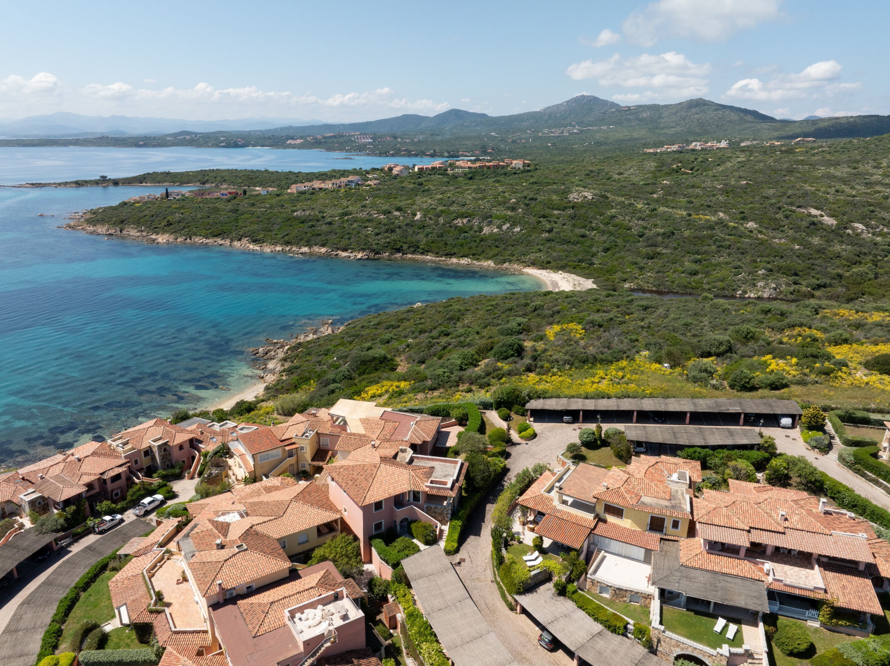 A coastal residential area with terracotta-roofed houses, a beach, and a mountainous backdrop.