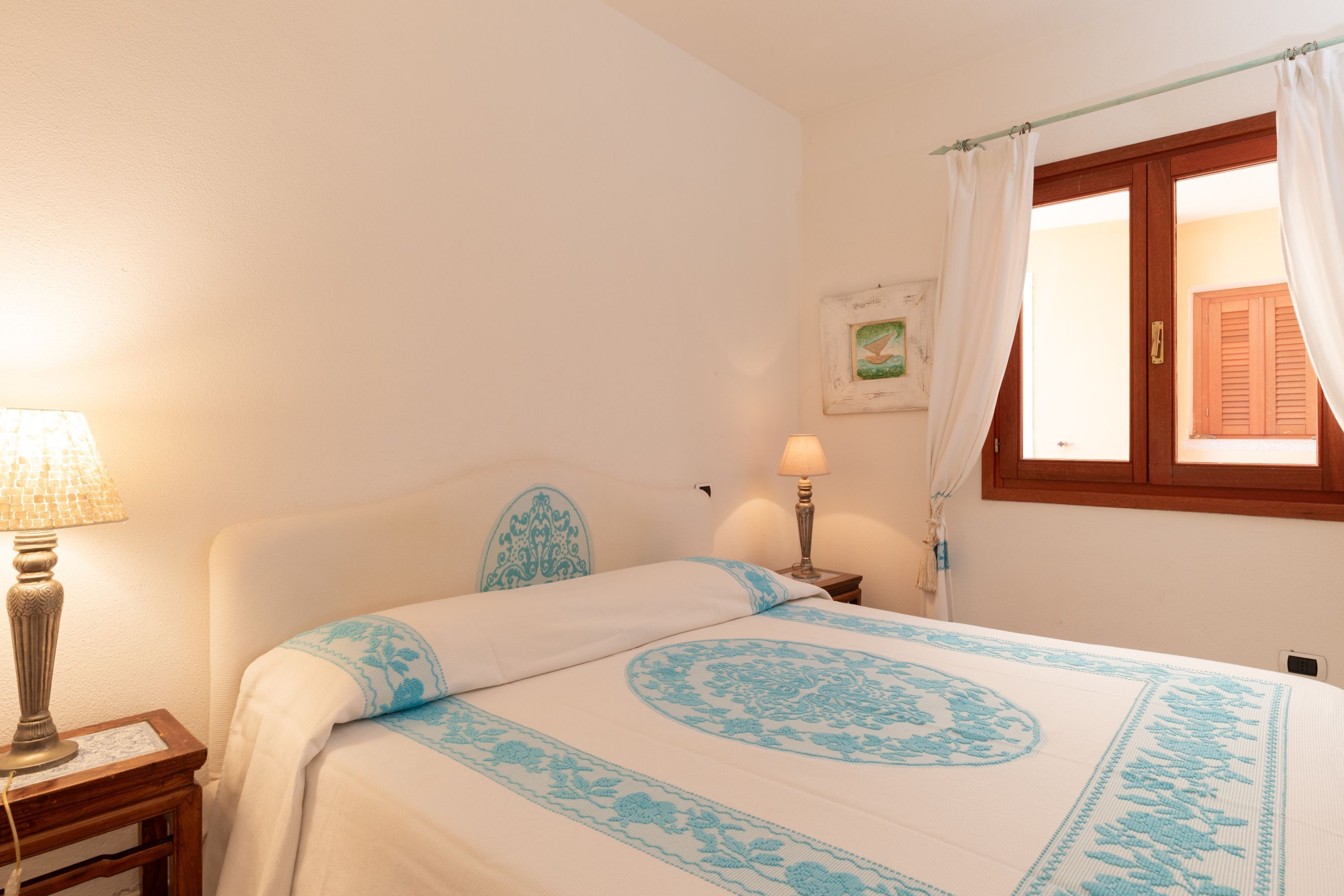 A bedroom with a bed, two bedside lamps, a window with curtains, and a picture on the wall.