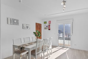 Un coin repas avec une table et des chaises blanches, une plante en pot et une vue à travers une porte vitrée.