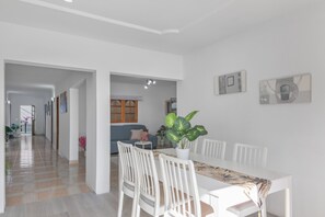 Un coin repas avec une table et des chaises blanches, une plante en pot et un couloir menant au salon.