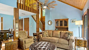 A spacious living room with a high ceiling, a wooden staircase, a large leather sofa, a tufted ottoman, and a dining area with wooden furniture. - Pet Friendly, Private Wooded Setting - Travelers Rest by Carolina Properties (Rutherfordton)