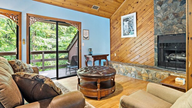 A cozy living room with a stone fireplace, leather ottoman, and a view of the outdoors. - Pet Friendly, Private Wooded Setting - Travelers Rest by Carolina Properties (Rutherfordton)