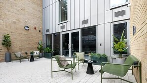 Un espace extérieur aménagé avec des chaises et des tables en métal vert, entouré de plantes en pot et d’un mur de briques.
