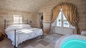 A bedroom with a stone wall, a bed with a white bedspread, a window with curtains, and a bathtub.