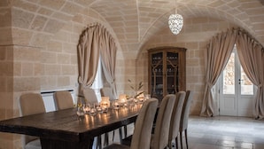A dining area with a long table set for six, surrounded by chairs, with a stone wall and arched ceiling.