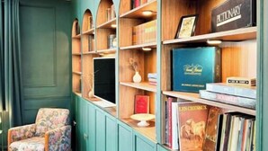 Un rincón acogedor para leer con un sillón estampado, una estantería llena de libros y objetos decorativos, y una mesita con un florero.