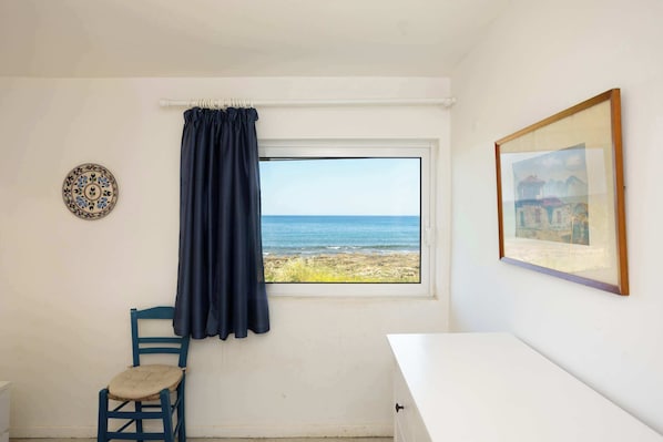 A room with a view of the sea, a chair, a table, and a framed picture on the wall.