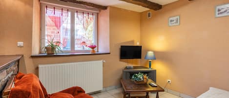 Una habitación acogedora con una silla de madera, una mesita con una lámpara, un televisor y una ventana con cortinas traslúcidas.