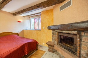 A bedroom with a bed, a window with curtains, a fireplace, and a wooden ceiling beam. - Holiday Home 'La Charrue' with Mountain View, Shared Pool and Wi-Fi (Villecomtal)