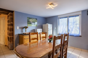 A dining area with a wooden table, chairs, a refrigerator, and a window with curtains. - Holiday Home 'La Charrue' with Mountain View, Shared Pool and Wi-Fi (Villecomtal)