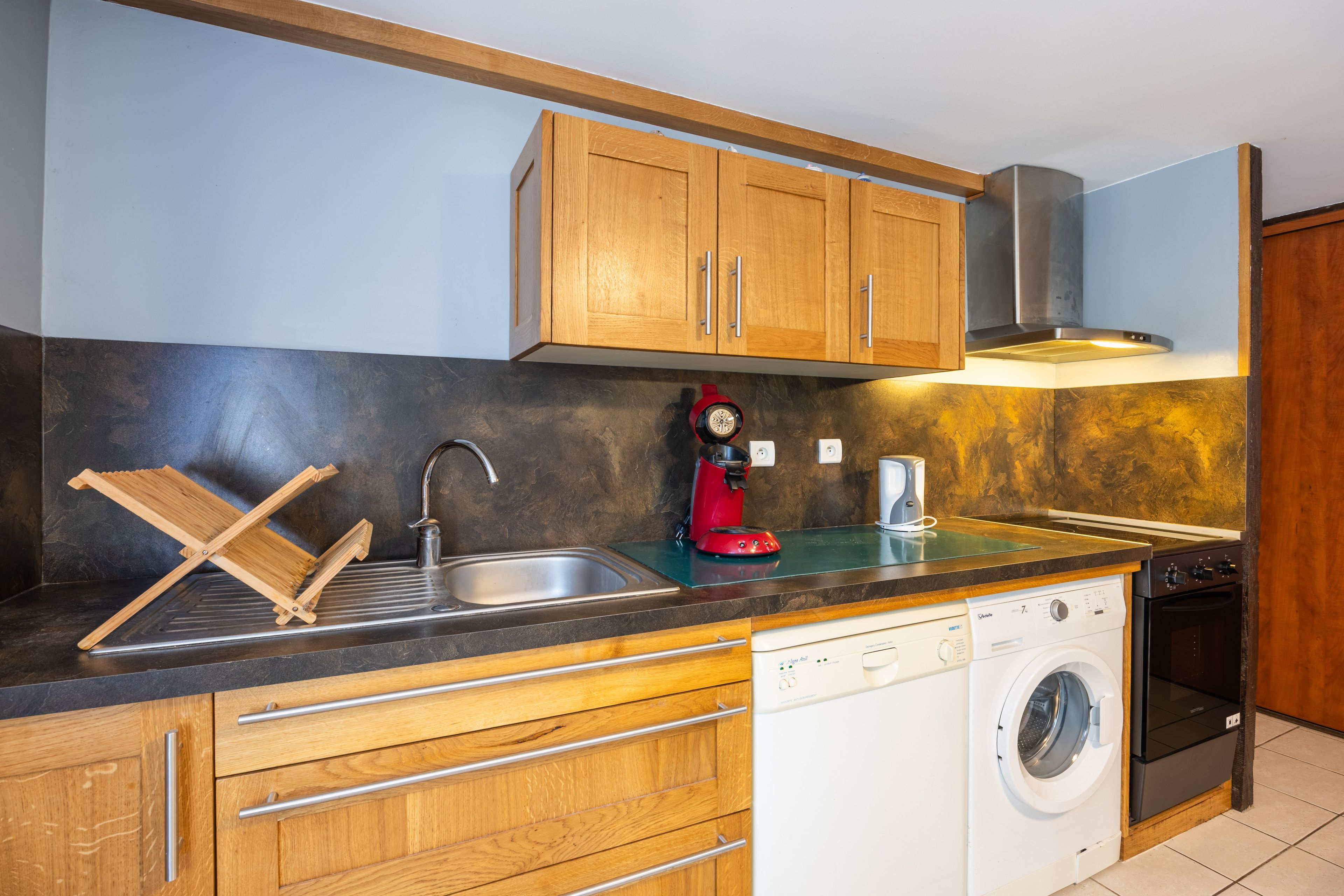 Una cocina moderna con armarios de madera, un lavamanos de acero inoxidable, una cafetera roja y un lavavajillas empotrado.