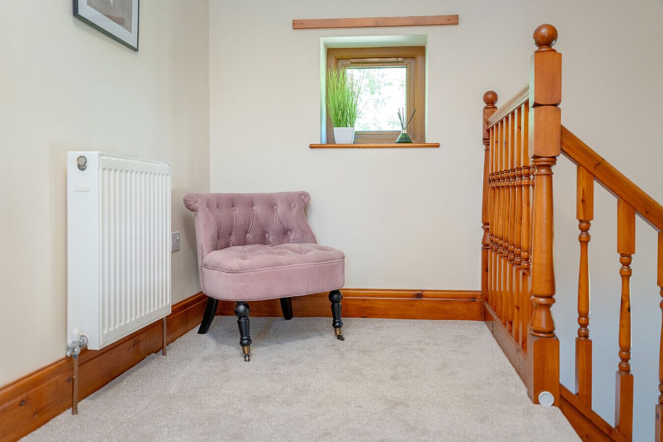 A corner room with a radiator, a pink tufted chair, a wooden staircase, and a small window with a plant.