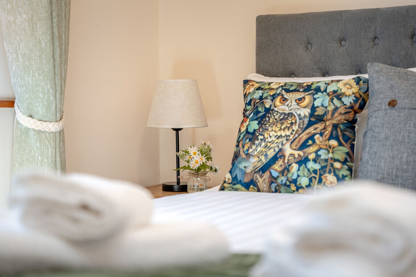 Een net opgemaakt bed met gerolde witte linnen, een nachtkastje met een lamp en een vaas met bloemen, en een kussen met een uilenprint.