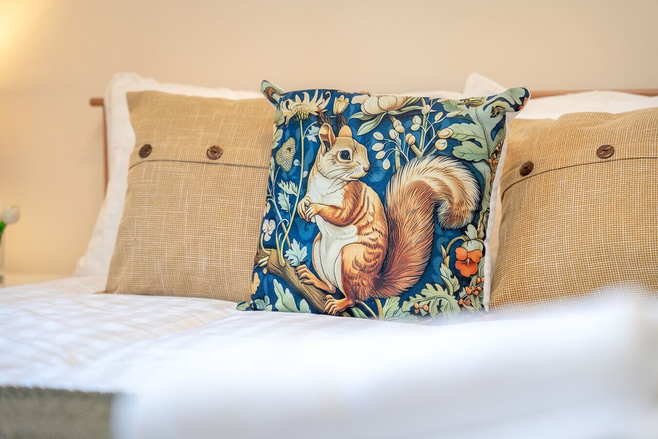 Una cama con colcha blanca, dos almohadas color beige y una almohada decorativa con diseño de ardilla.