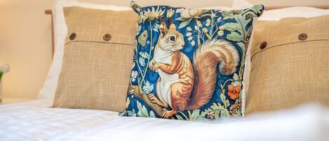 Una cama con colcha blanca, dos almohadas color beige y una almohada decorativa con diseño de ardilla.