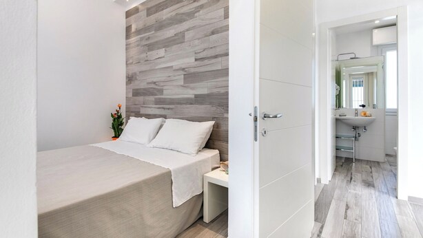 Ett modernt sovrum med en vägg som har en träpanel som accent, en säng med vita sängkläder, ett sängbord med en växt och utsikt över badrummet med handfat och spegel.