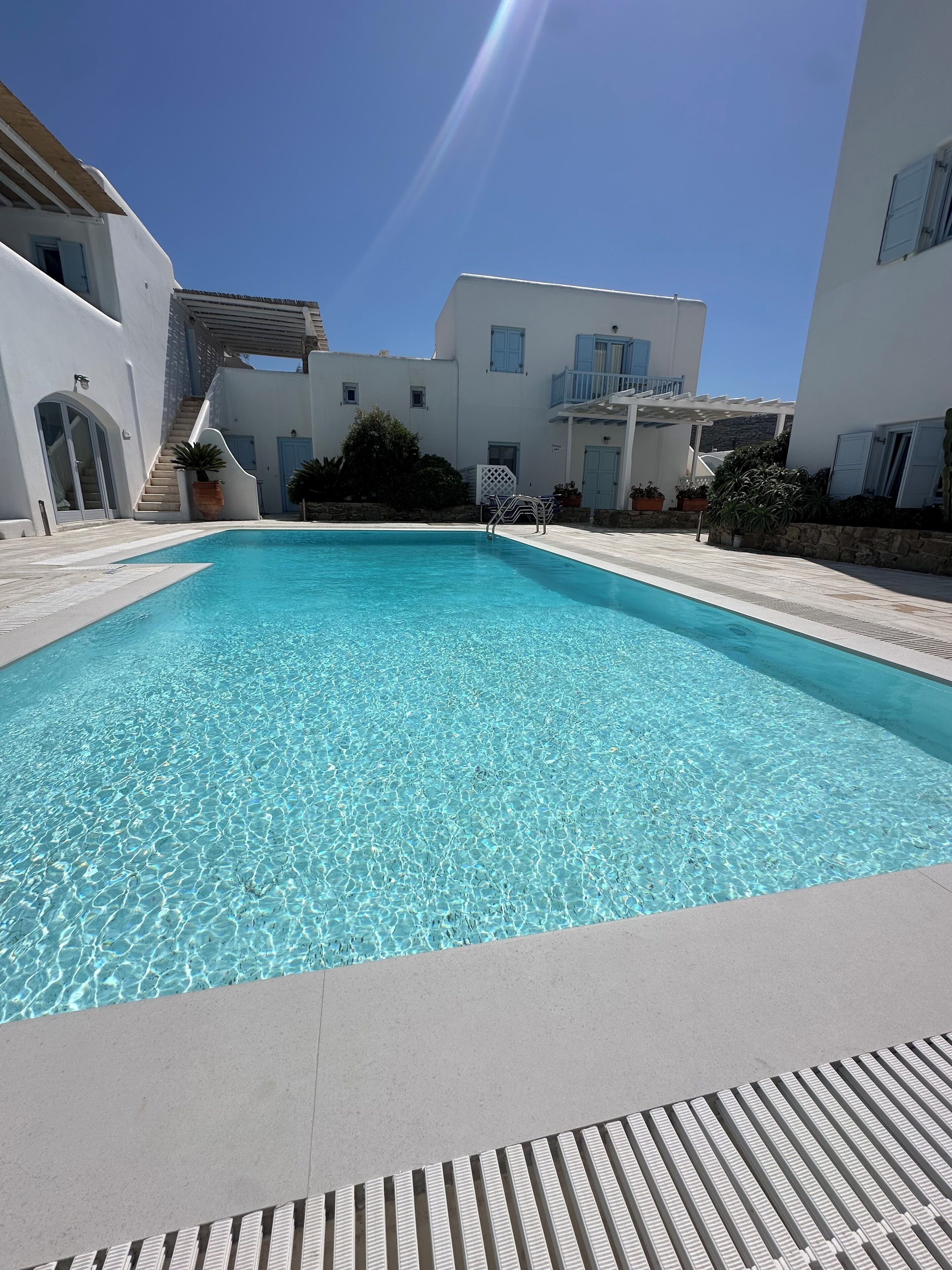 Una piscina rectangular con agua azul cristalina, rodeada por una terraza de baldosas y edificios blancos con balcones.