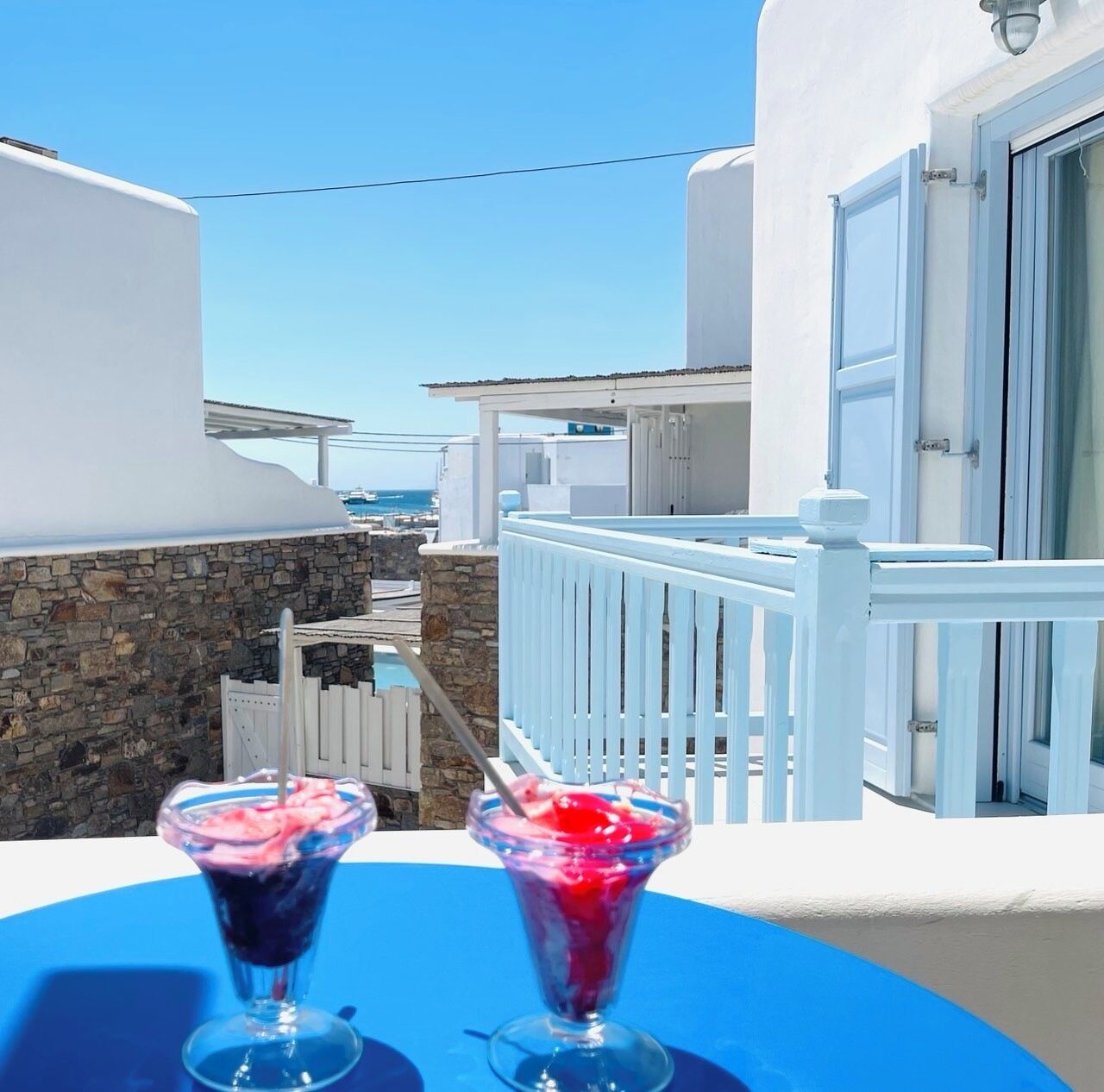 A balcony with two drinks on a blue table, overlooking a pool and buildings with white and blue architecture.
