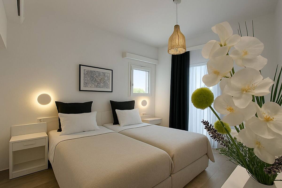 A modern hotel room with a double bed, bedside tables, a hanging light fixture, a window with curtains, and a framed artwork on the wall.