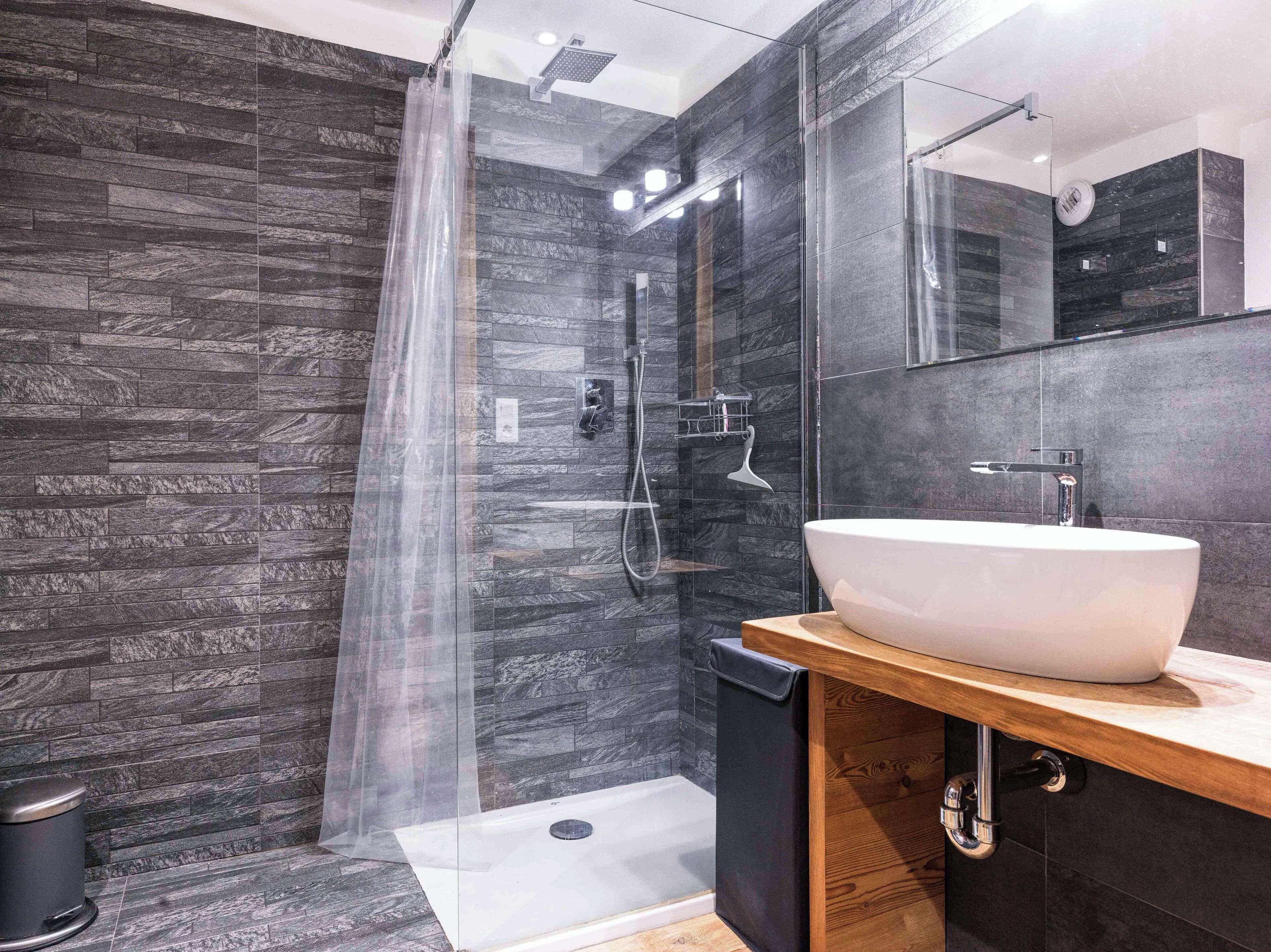 A modern bathroom with a large white sink, a glass shower enclosure, and dark stone walls.